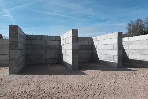 Budowa muru oporowego z bloczków betonowych. Konstrukcja tworzy zarys pomieszczeń na piaszczystym placu budowy.